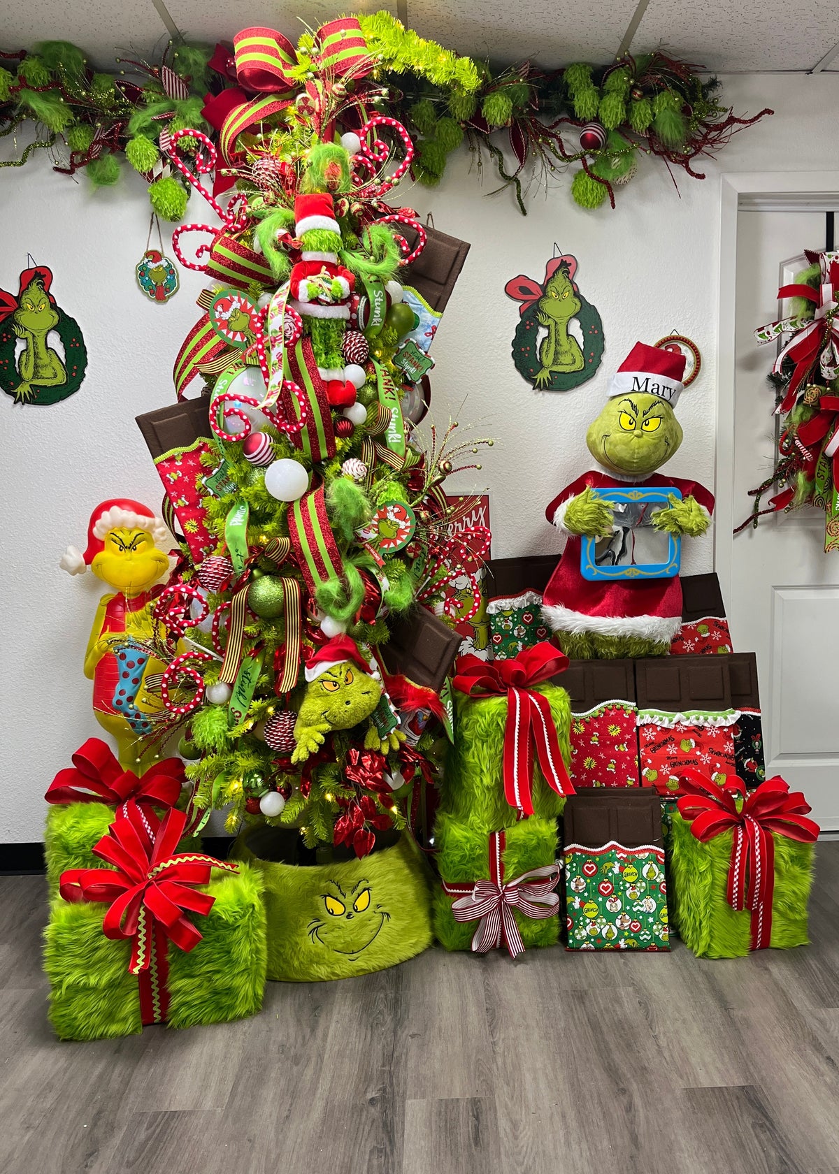 A festive Grinch-themed holiday display featuring a decorated Christmas tree with green ornaments, red ribbons, Grinch figurines, wrapped presents, and holiday wall decorations in an indoor room.